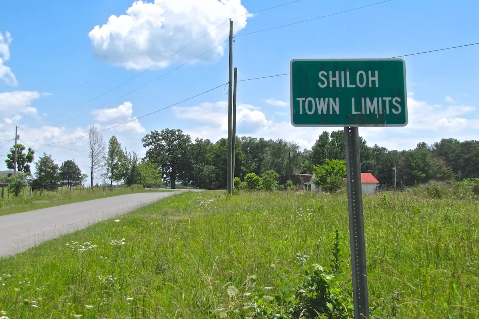 Green sign with white text 'Shiloh Town Limits' against green grassy field with small road on left.